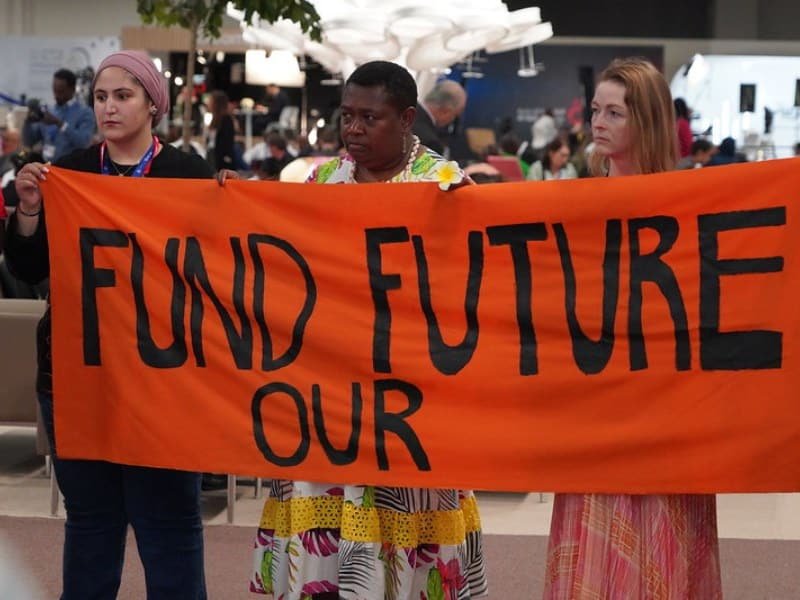 tres personas con una bandera que dice "financiá nuestro futuro" en la COP28.